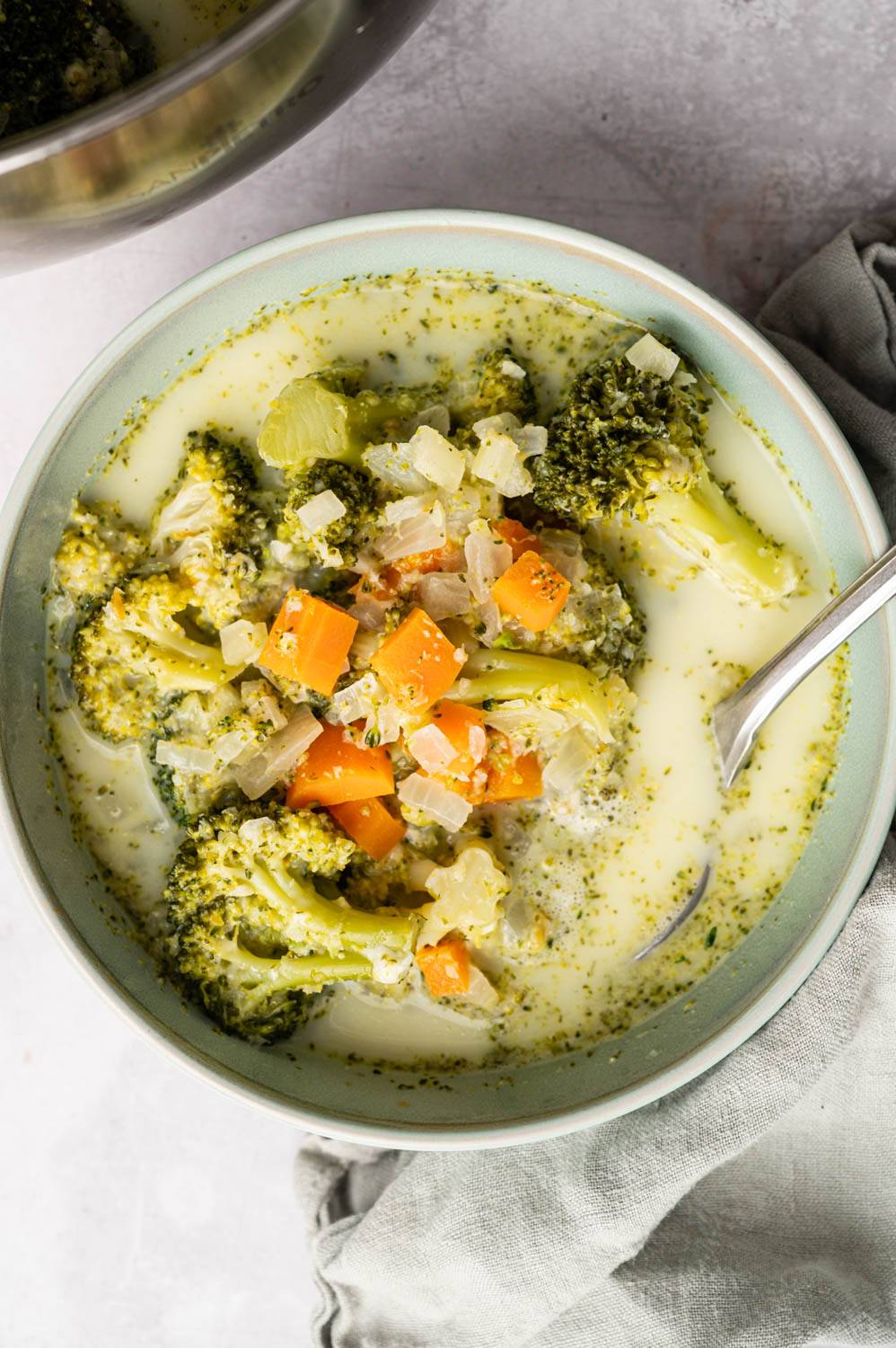 A bowl of creamy broccoli soup with visible broccoli florets, diced carrots and onions. The soup looks warm and cozy with a spoon inside. A light gray napkin lies on a textured surface next to the bowl.