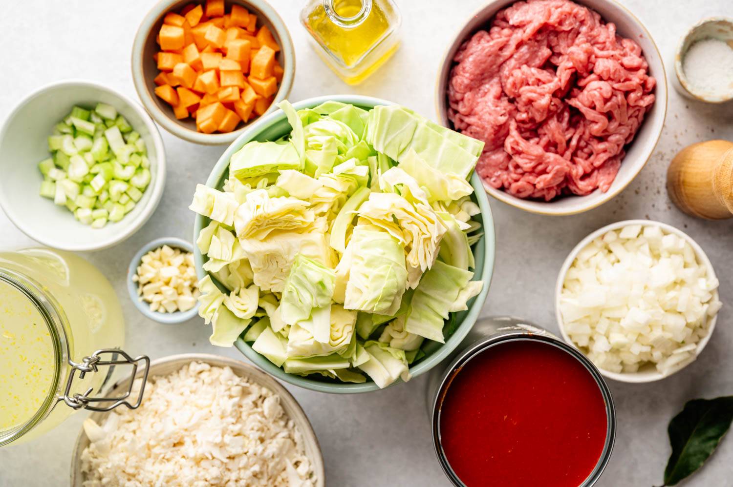 Assorted fresh ingredients including chopped cabbage, minced meat, diced onions, carrots, celery, rice, minced garlic, tomato paste and a can of broth on a white surface. A small flask of oil and a salt bowl can also be seen, indicating that a meal is being prepared.