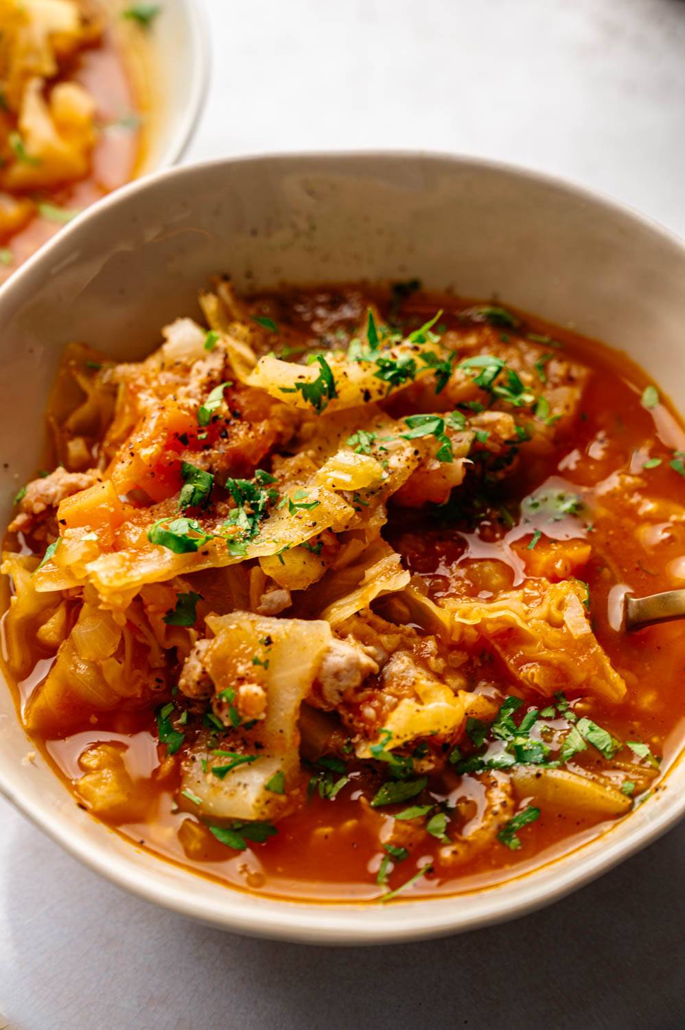 Close-up of bowl filled with hearty cabbage soup with bright orange broth, tender cabbage, meat cubes, and garnished with fresh parsley.