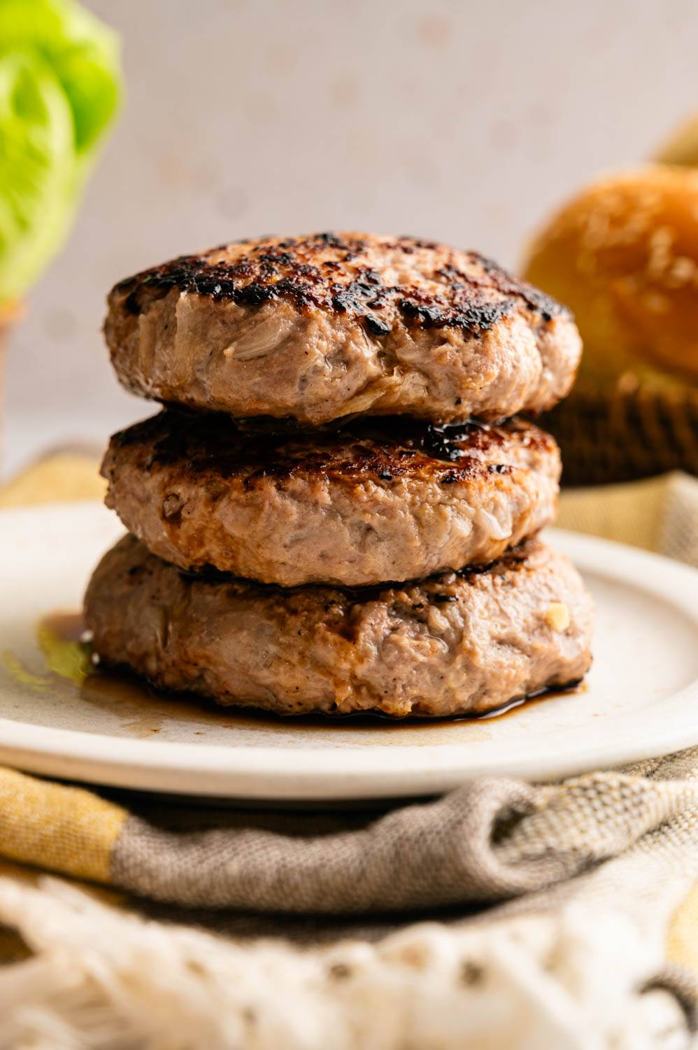 Three grilled turkey burger patties stacked on a white plate, charred and juicy, with a sesame bun and lettuce blurred in the background.