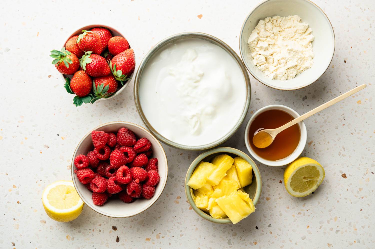 Ingredients for high-protein Greek yogurt popsicles arranged on a countertop, including Greek yogurt, protein powder, honey, lemon, strawberries, raspberries, pineapple, and fresh lemon halves.