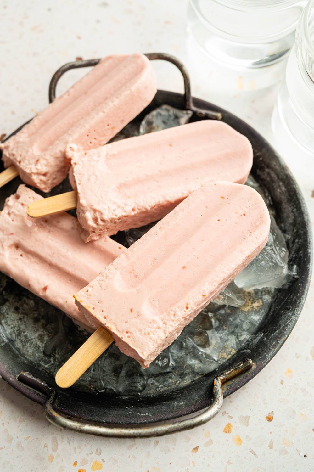 Creamy high-protein Greek yogurt popsicles served over ice in a rustic metal dish with glasses of water in the background.
