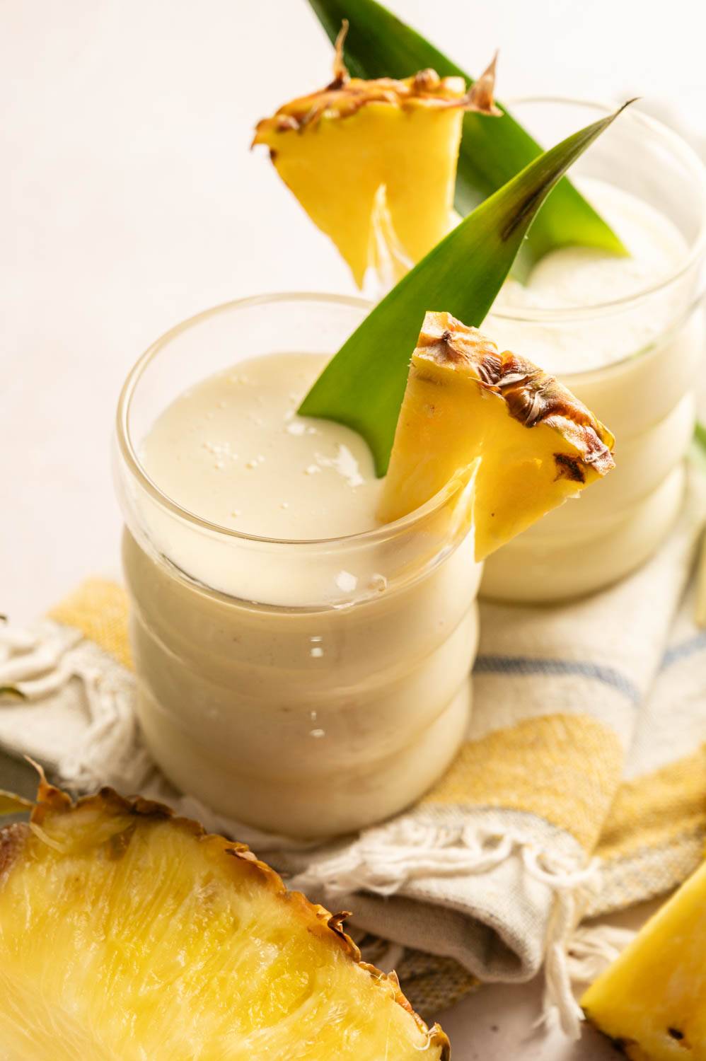 Vertical close-up of a creamy piña colada protein smoothie in a glass, garnished with a fresh pineapple wedge and pineapple leaf.
