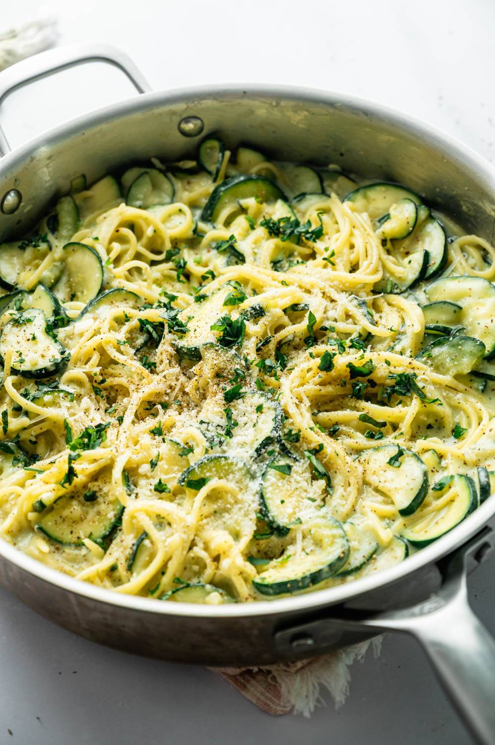 Flat lay of raw ingredients for zucchini pasta on a white marble surface, including dry linguine, two whole zucchini, a bowl of cottage cheese, pesto, minced garlic, bone broth, shredded Parmesan, fresh basil, and a lemon.