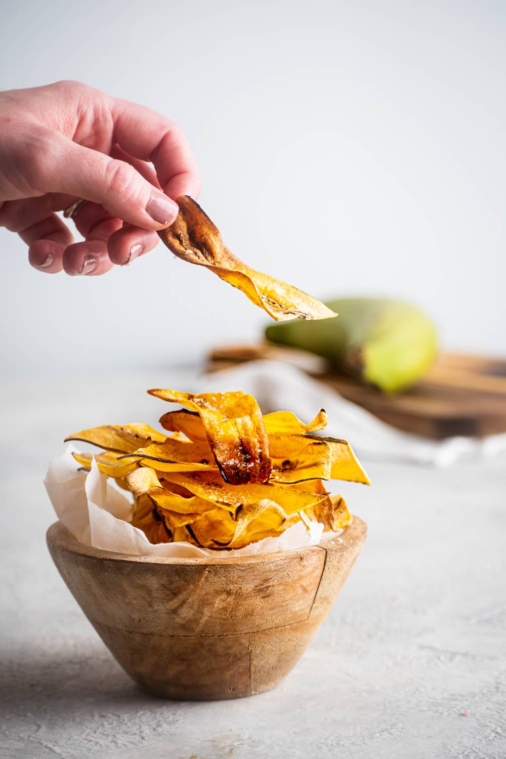Holding crisp homemade plantain chips in hand, topped with a rustic wooden bowl filled with golden fries and a green plantain on the background.