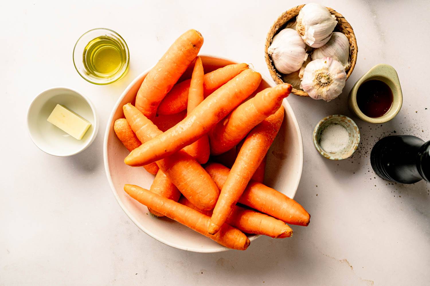 Ingredients for honey roasted carrots arranged on a countertop, including whole carrots, garlic, butter, olive oil, honey, salt, and black pepper.