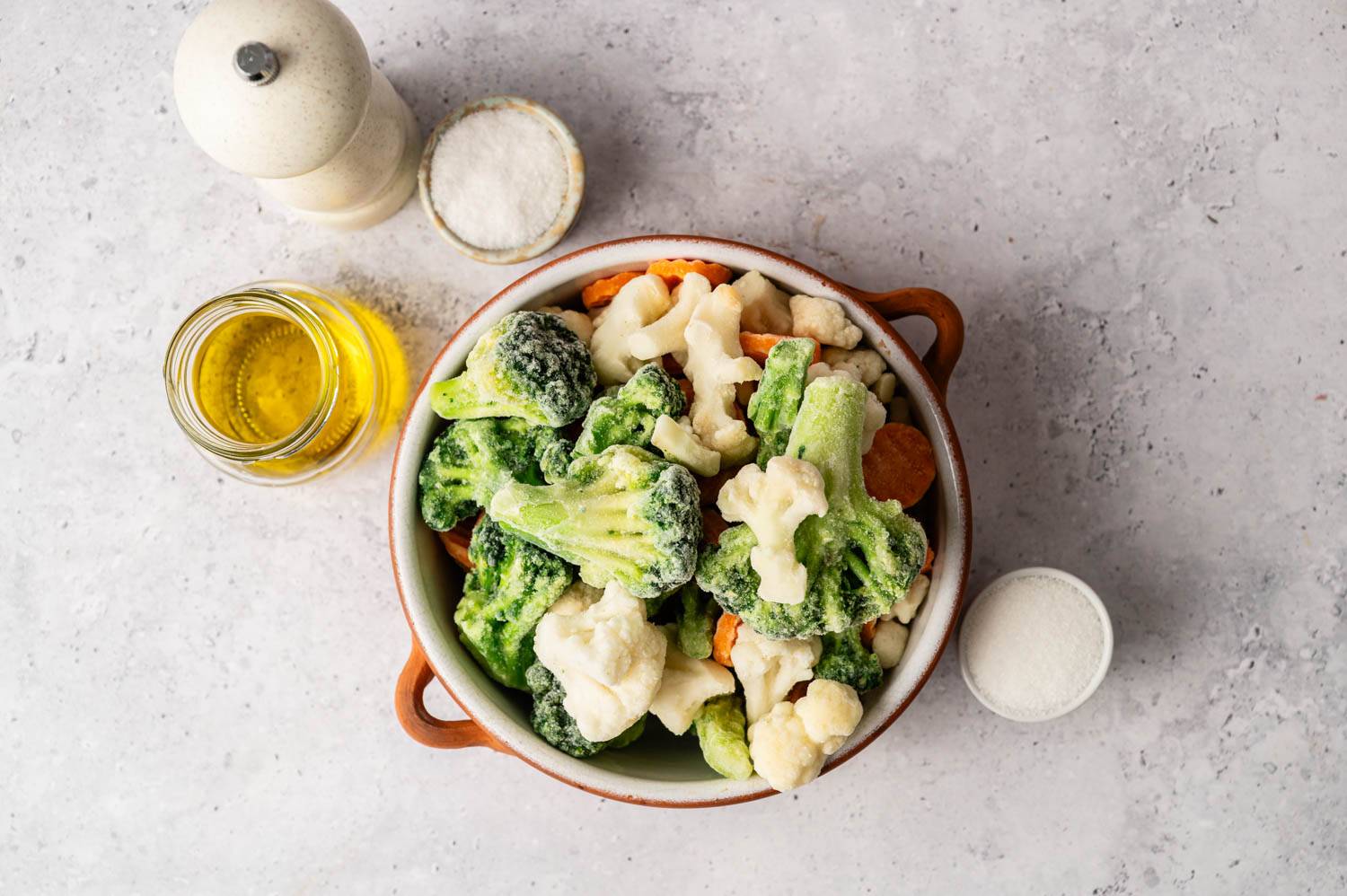 A bowl of frozen mixed vegetables, including broccoli and cauliflower, on a gray surface. Olive oil, a salt pan and a pepper grinder can be seen nearby.