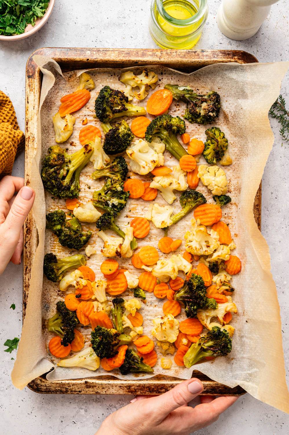 Arrange roasted broccoli, cauliflower, and carrot slices on a baking sheet on parchment paper. The tray is held in hand with olive oil and seasonings beside it, conveying a sense of freshness.