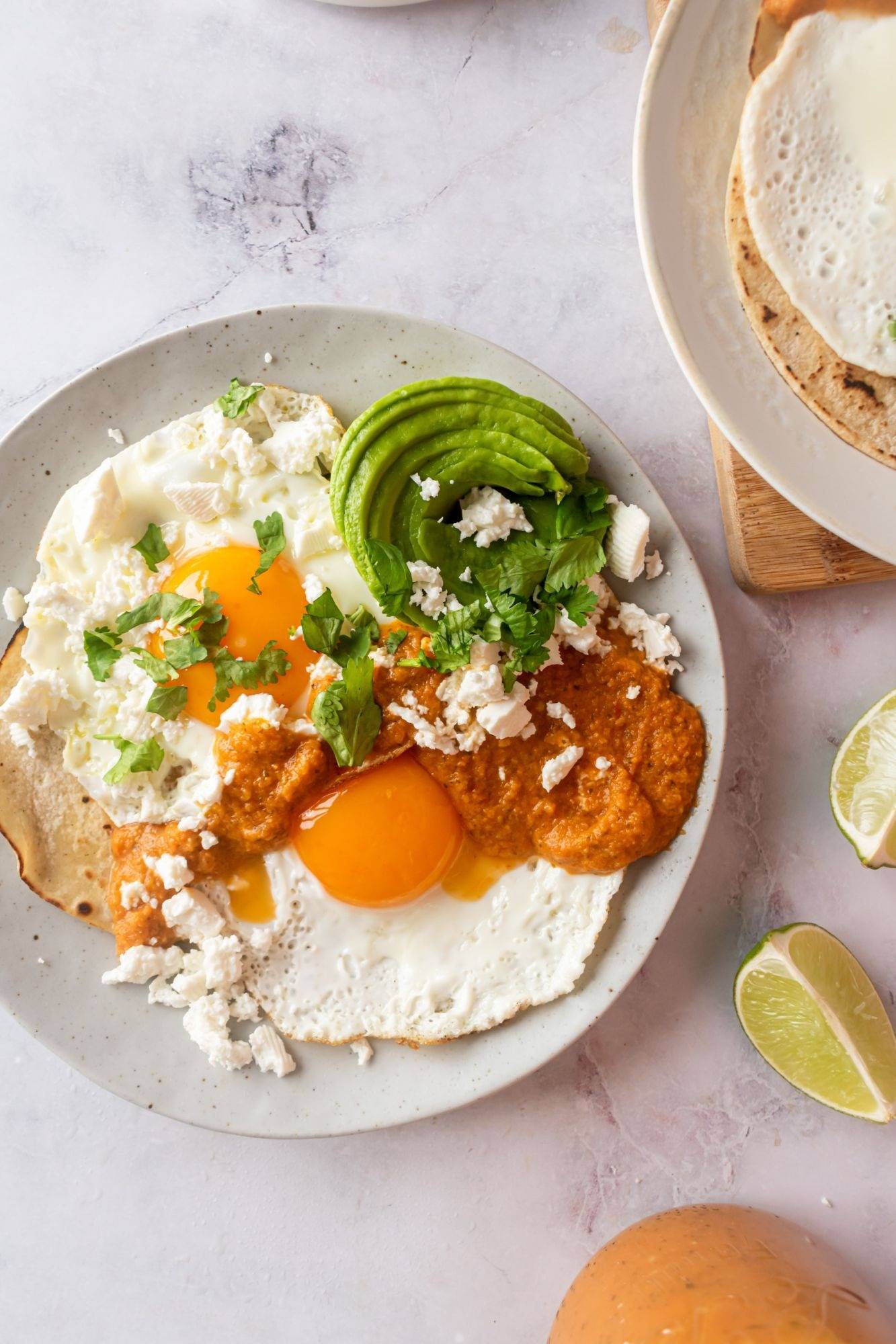 Wooden boarded Huevos Rancheros has eggs beside the sun on the tortilla, topped with red sauce, thinly sliced avocado, crumpled Queso Fresco and Fresh Cilantro with lime wedges on the sides.  