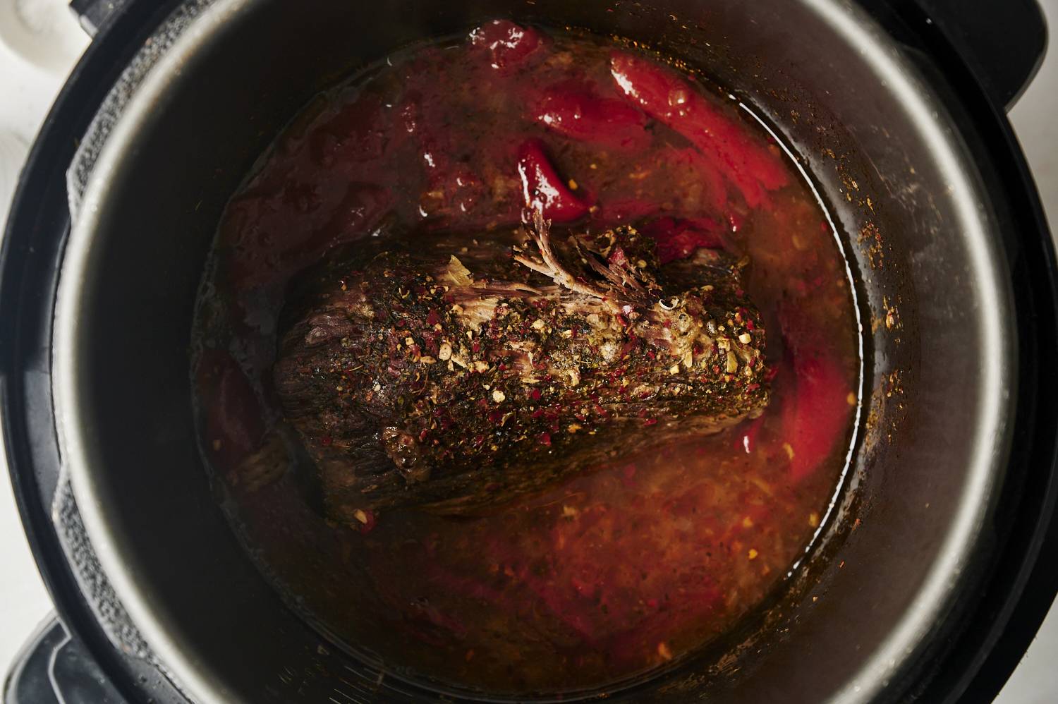 A cooked beef roast in an Instant Pot surrounded by a savory broth with red peppers, herbs, and spices, showing the meat tender and ready for shredding.