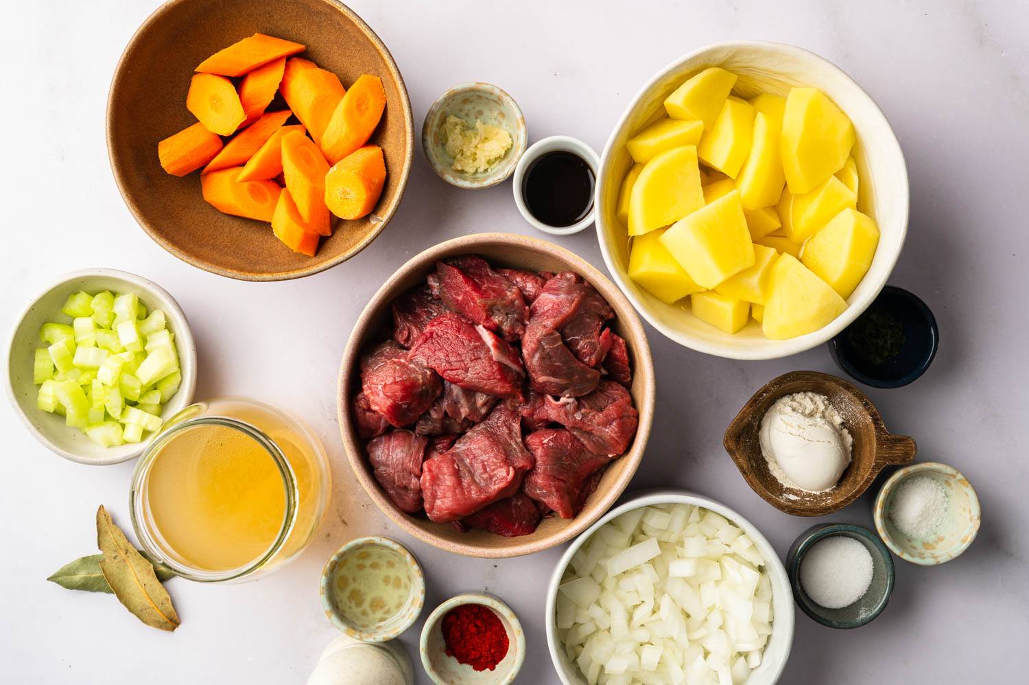 A variety of fresh ingredients for cooking: sliced carrots, potatoes, celery, diced onions, cubed beef, broth, and seasonings.