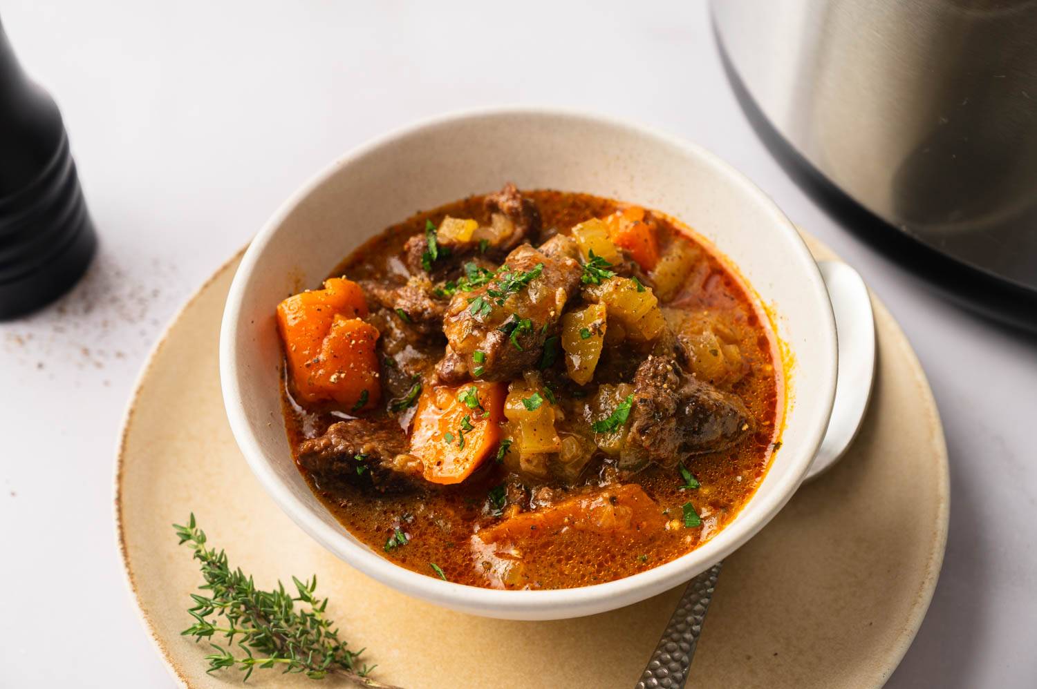 A bowl of hearty beef stew with carrots and potatoes, garnished with parsley, on a cream-colored plate. A pepper grinder is nearby.