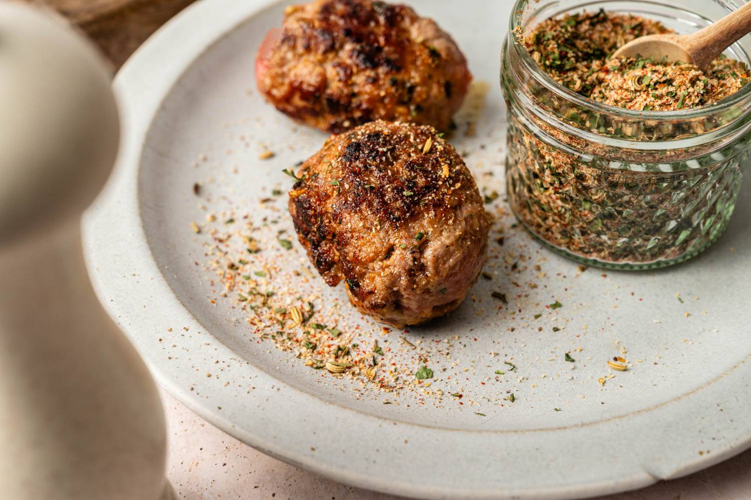 Two brown meatballs are placed on a white plate and sprinkled with herbs. A jar of seasonings and a small wooden spoon sit on the side for a delicious homemade feel.