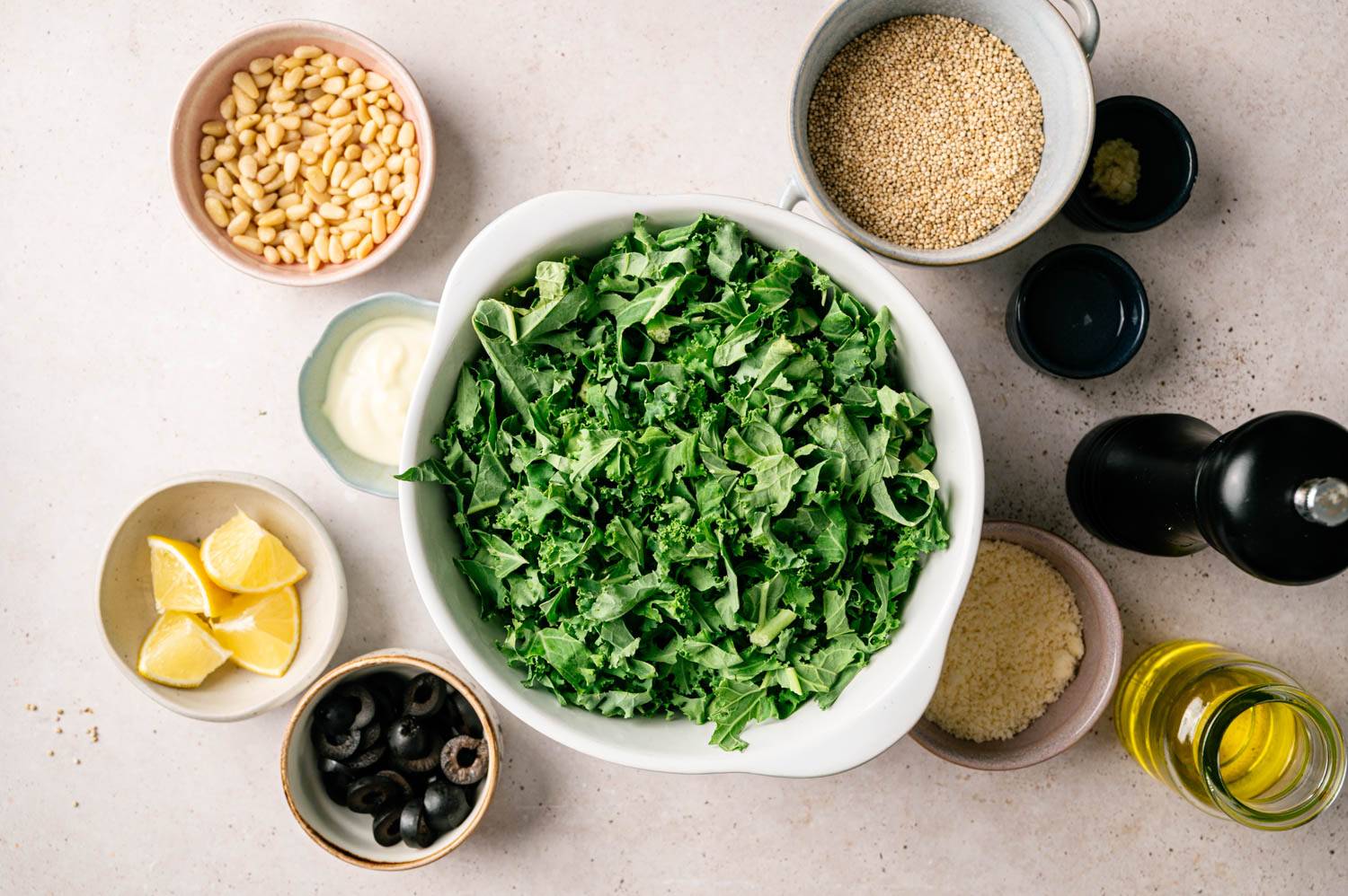 A white bowl of fresh kale is surrounded by ingredients: pine nuts, lemon slices, black olives, quinoa, parmesan cheese, oil and creamy dressing for a refreshing, healthy vibe.