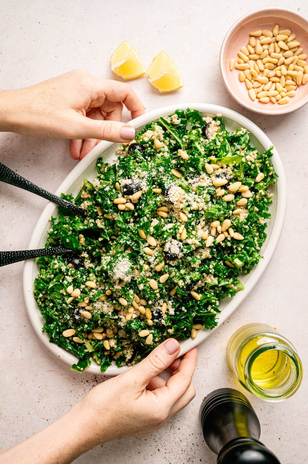 Fresh kale salad with pine nuts and grated cheese on an oval plate. Nearby are two lemon slices, a bowl of pine nuts and olive oil. Use both hands to adjust the plate.