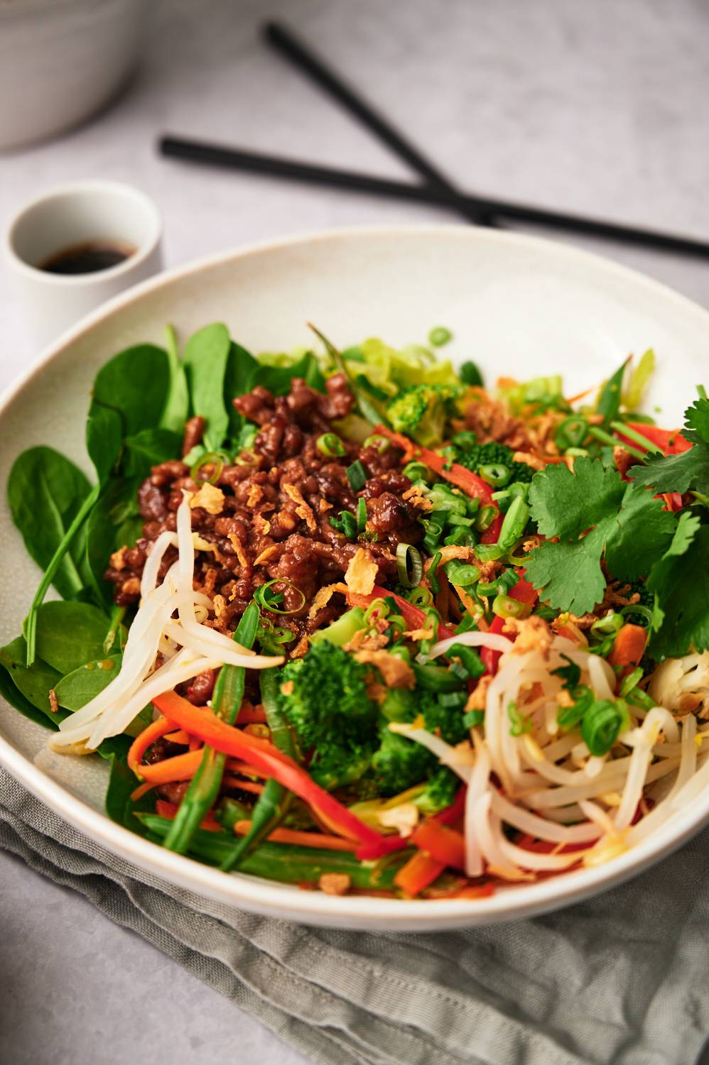A bowl of vibrant mixed salad that includes spinach, broccoli, carrots, bean sprouts and grated meat, topped with herbs and green onions.