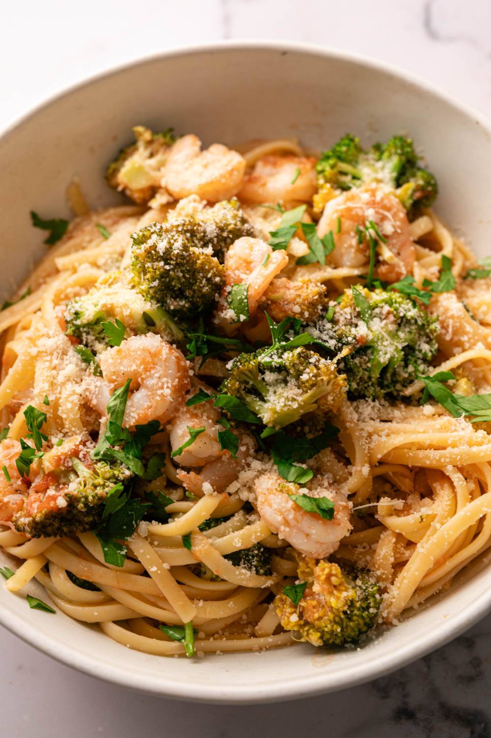 A bowl of linguine pasta topped with shrimp, broccoli, and parsley, sprinkled with Parmesan cheese.