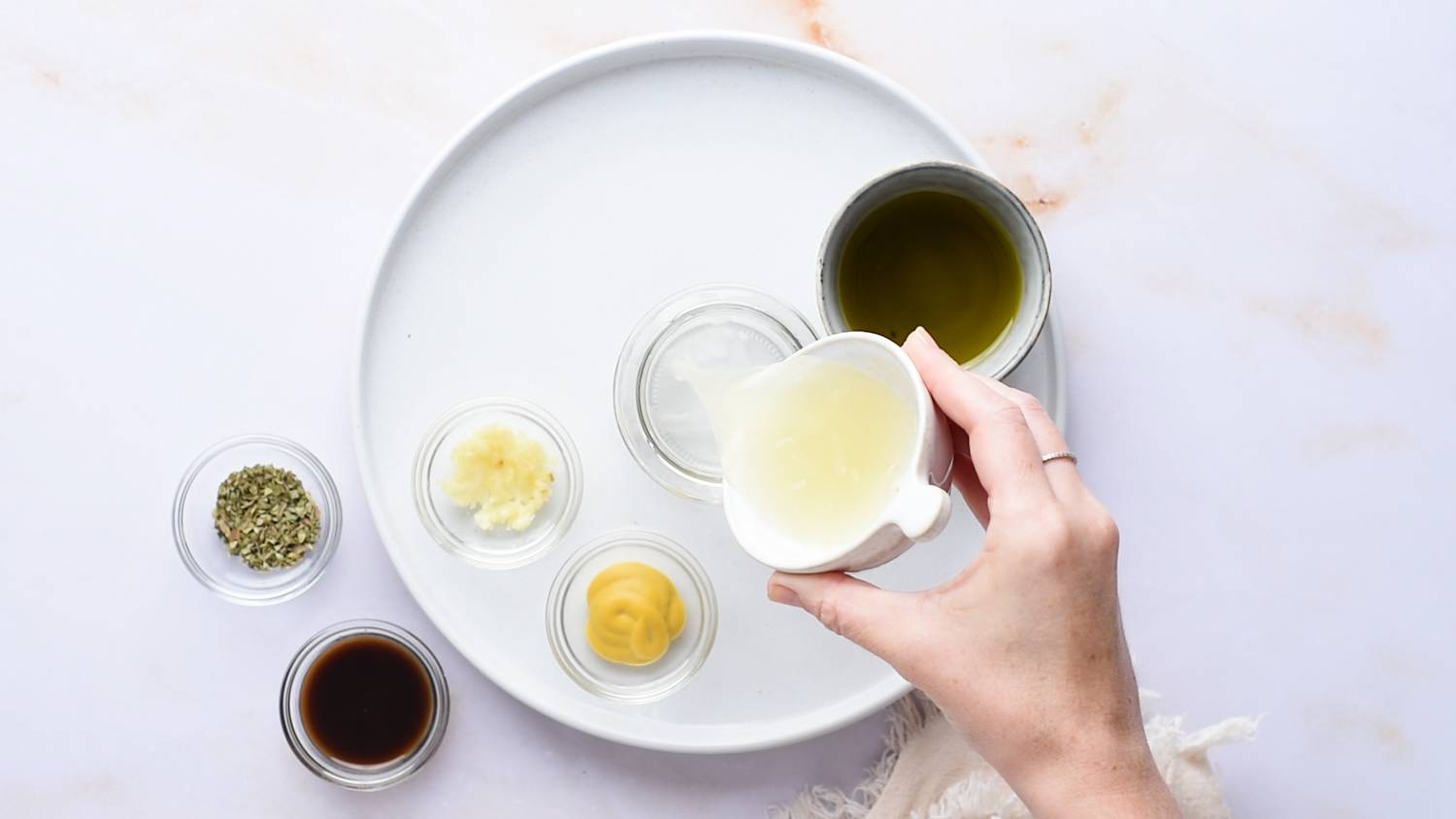 Top view of white plate with small glass bowls containing various ingredients including herbs, mustard and oil. Pour the lemon juice into the bowl with one hand.