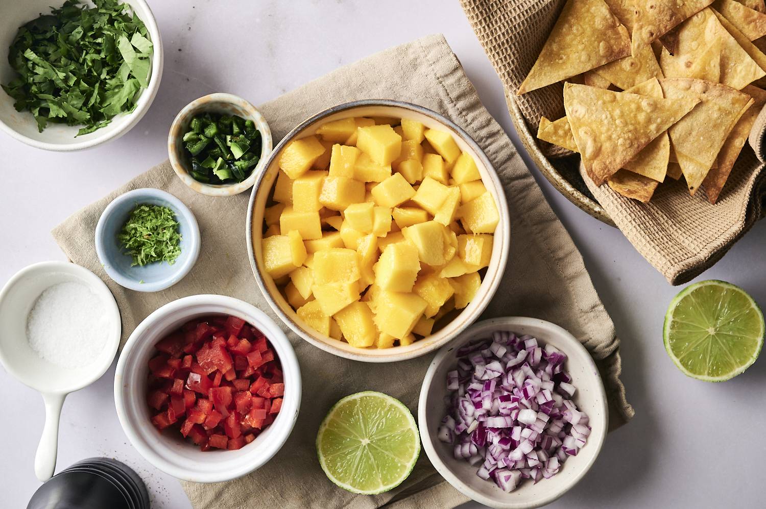 Elevated landscape with fresh mango salsa ingredients in the bowl, including diced mangoes, red onions, red bells, jalapeno, lime crust, coriander and a basket of crispy tortillas.