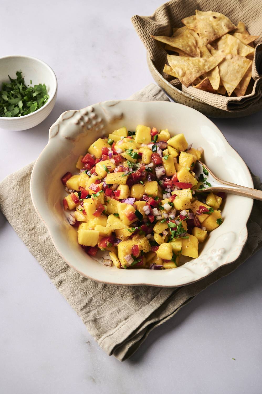 Fresh mango salsa (Salsa) in a white bowl with wooden spoons topped with mango, red onion, red bell pepper, jalapeno and cilantro served with tortilla fries.