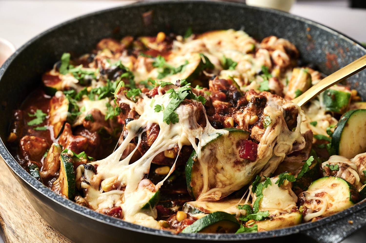 Close-up of cheesy vegetable and meat dishes with cilantro and bubbling in a frying pan with a golden spoon.