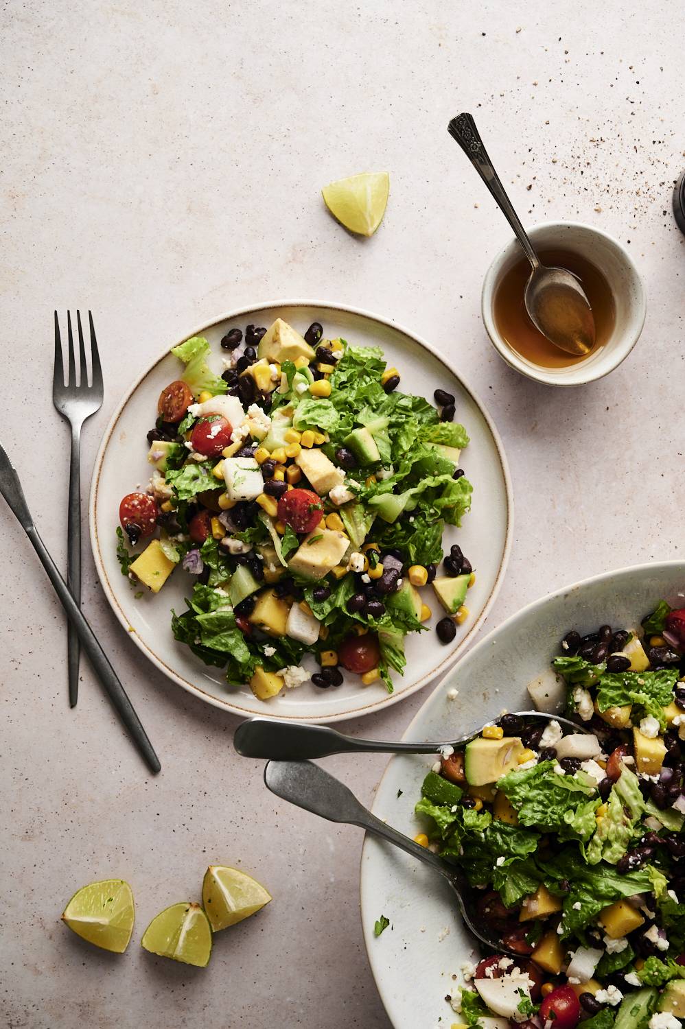 A vibrant Mexican chopped salad topped with fresh romaine, black beans, corn, mango, avocado, tomatoes and jicama, with lime wedges and a bowl of honey seasoning.
