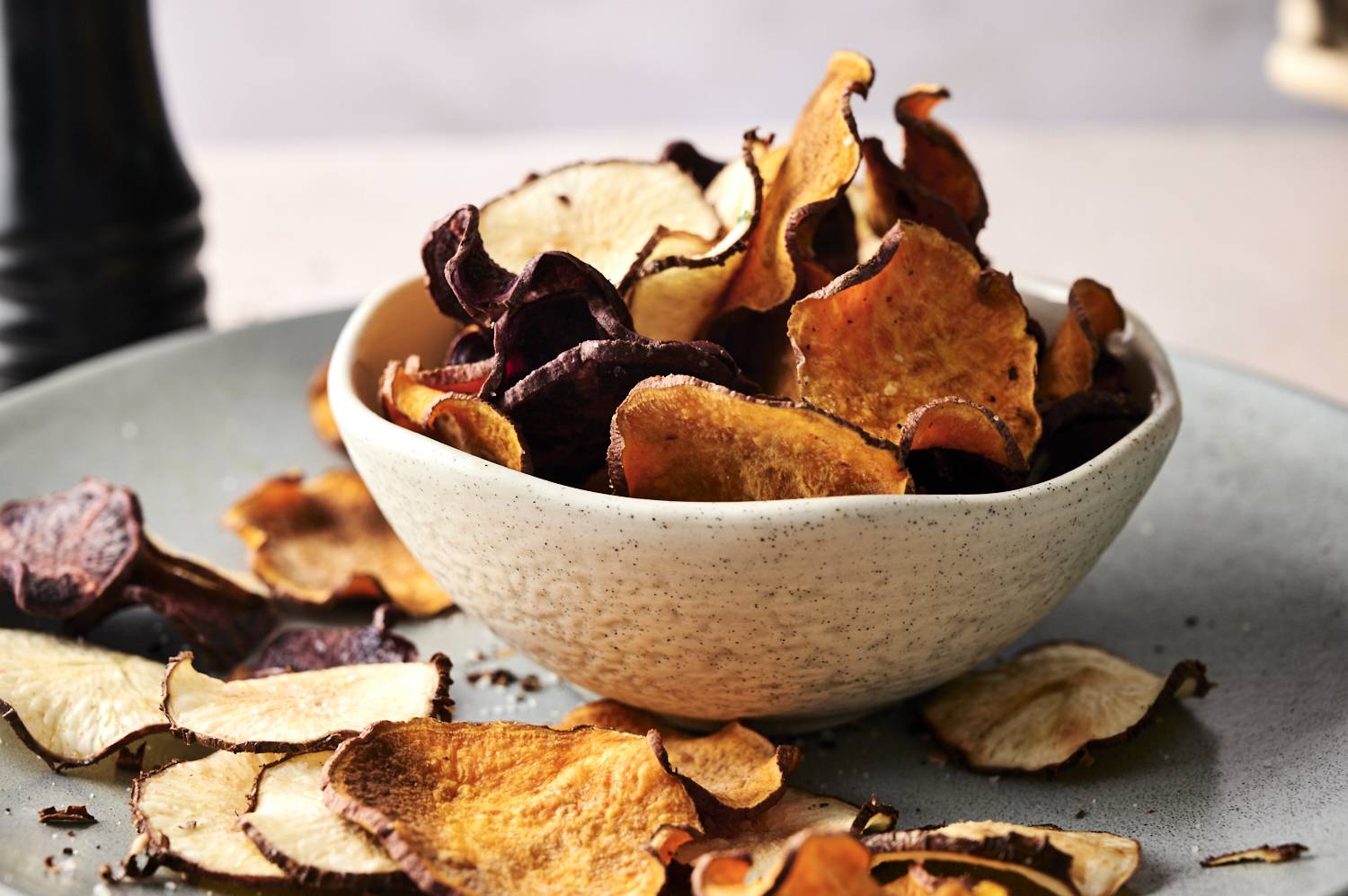 A bowl filled with colorful vegetable fries sits on a plate surrounded by scattered chips of various shapes and colors.