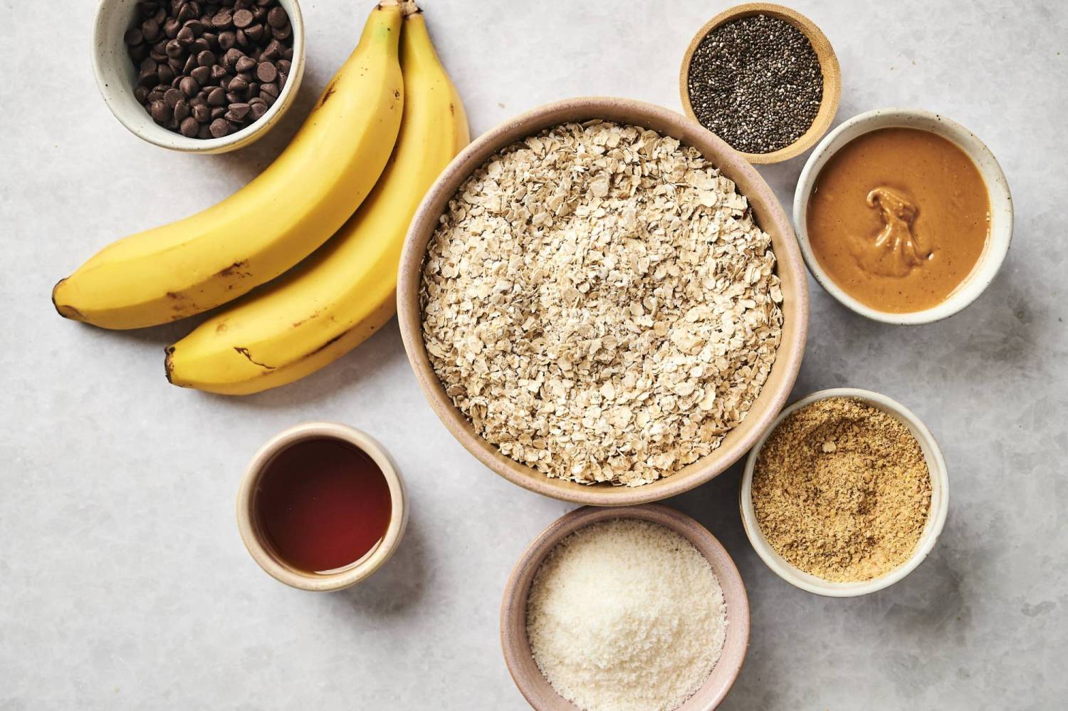 Flat ripe bananas, a bowl of oats, peanut butter, chocolate chips, chia seeds and sweetener on a light background.