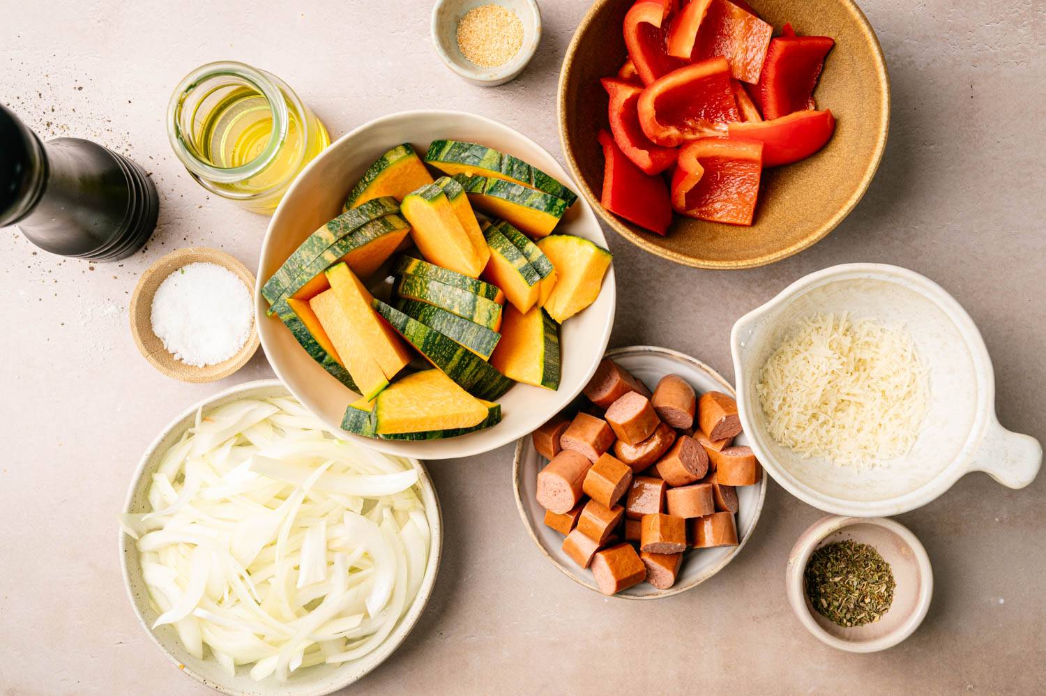 Ingredients on the table include chopped squash, red bell peppers, sliced ​​onions, sausage slices, shredded cheese and seasonings. There is a bottle of oil and salt nearby.