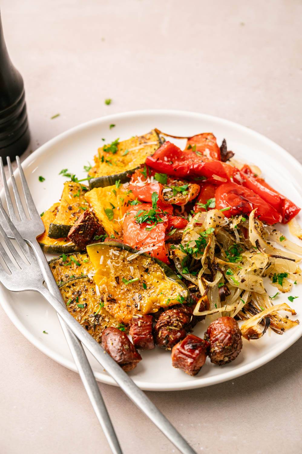 A plate of colorful grilled vegetables including zucchini, red peppers and onions, served with parsley. Serve with brown sausage slices. Two forks placed on the plate create an attractive and appetizing appearance.