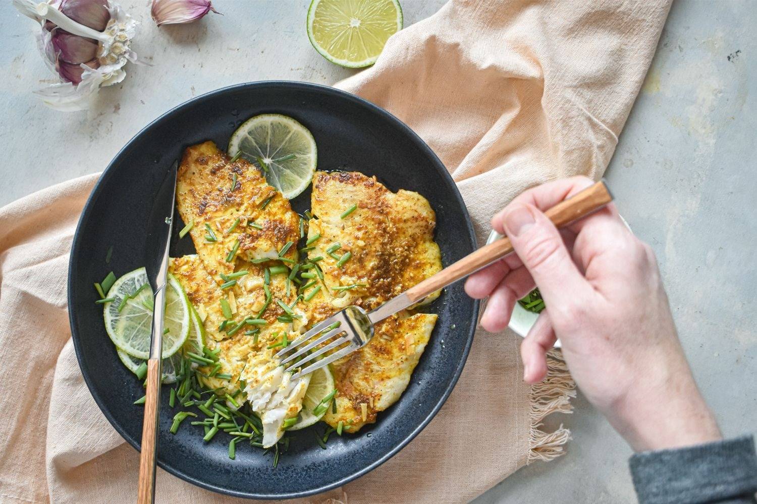 Garlic tilapia is pan-fried on a plate with butter and Old Bay seasoning using a fork and knife.