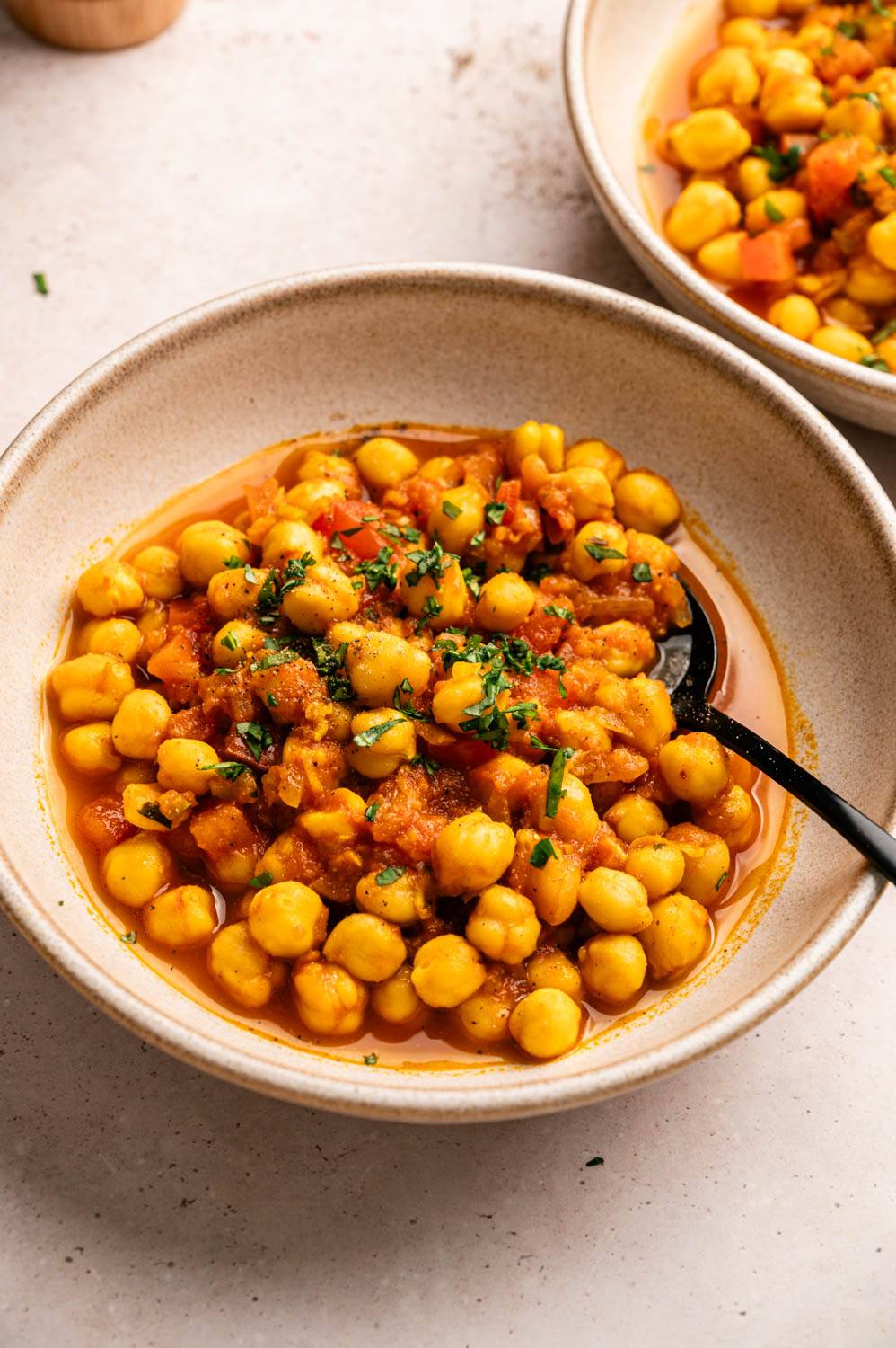 A bowl of vibrant chickpea stew garnished with fresh herbs, featuring golden chickpeas and diced tomatoes in a rich broth, alongside a black spoon.