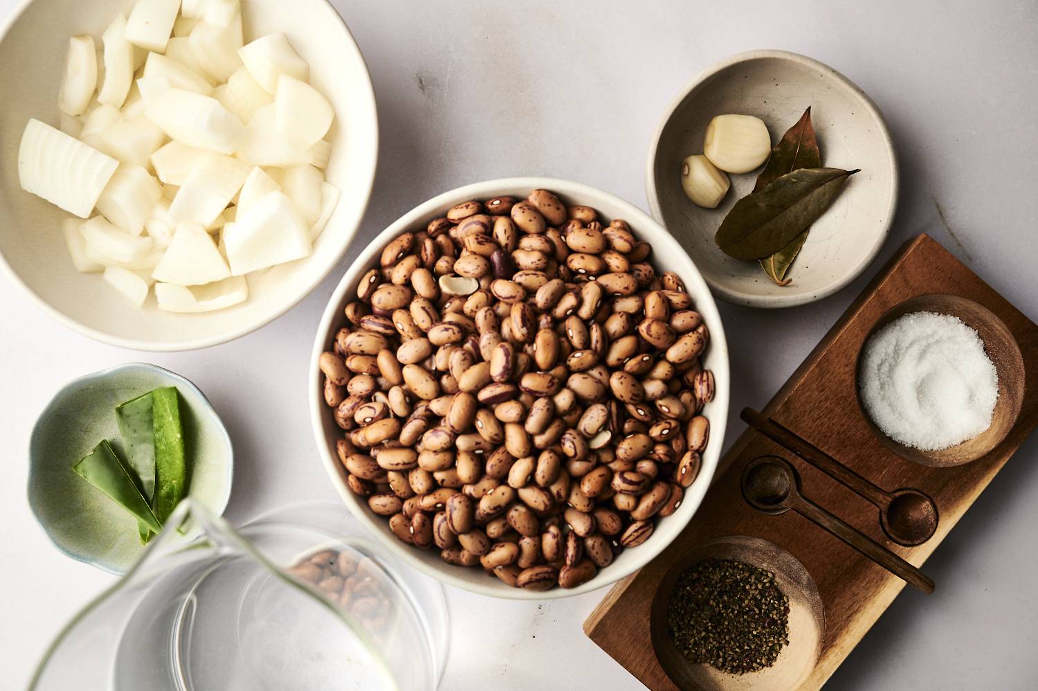 Ingredients for cooking: diced onions, pinto beans, garlic, bay leaves, salt and green greens on a light surface.
