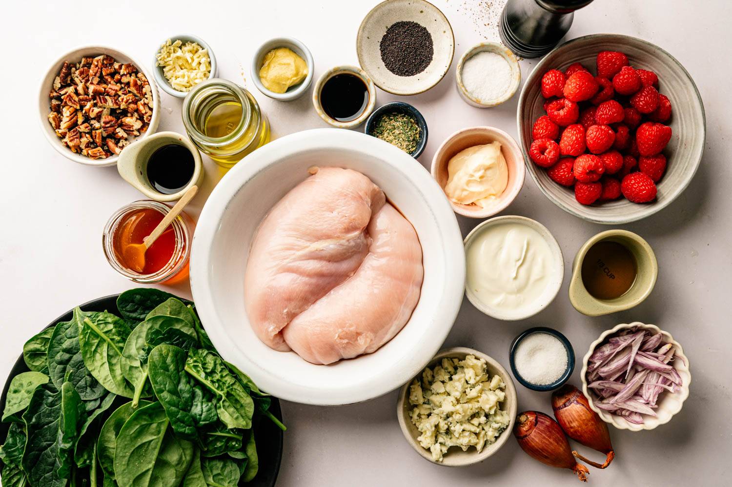 Two raw chicken breasts in a bowl are surrounded by ingredients like fresh spinach, raspberries, pecans, spices, oil, honey, and garlic on a white surface.