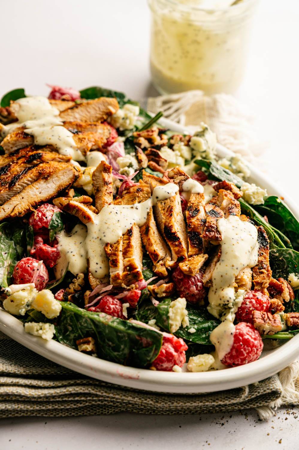 Grilled chicken salad with raspberries, spinach, pecans, and blue cheese crumbles, drizzled with creamy dressing on a white plate.