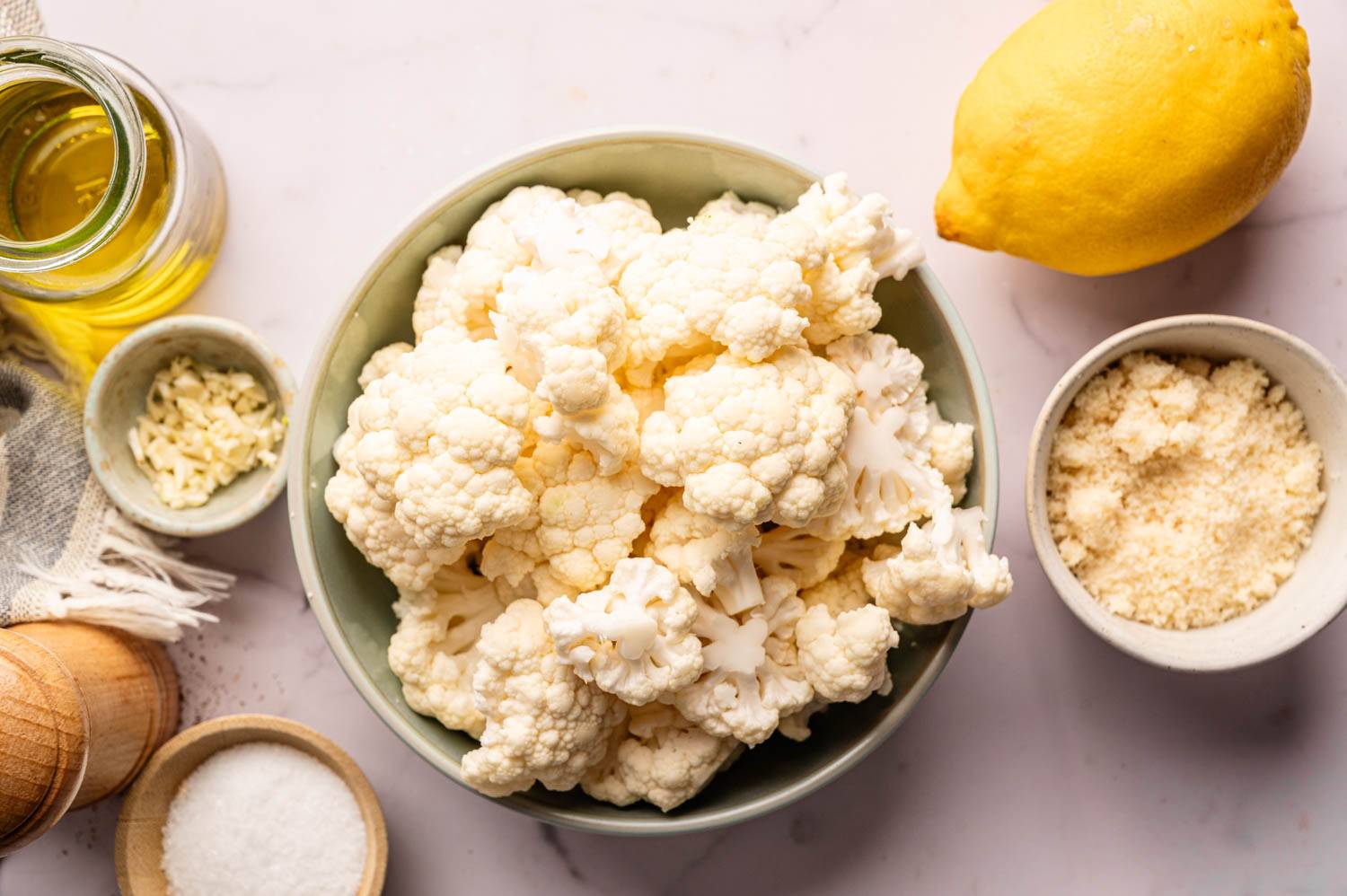 On a light marble surface lies a bowl of cauliflower, next to lemon, a jar of olive oil, minced garlic, a bowl of grated cheese, salt and pepper mill.