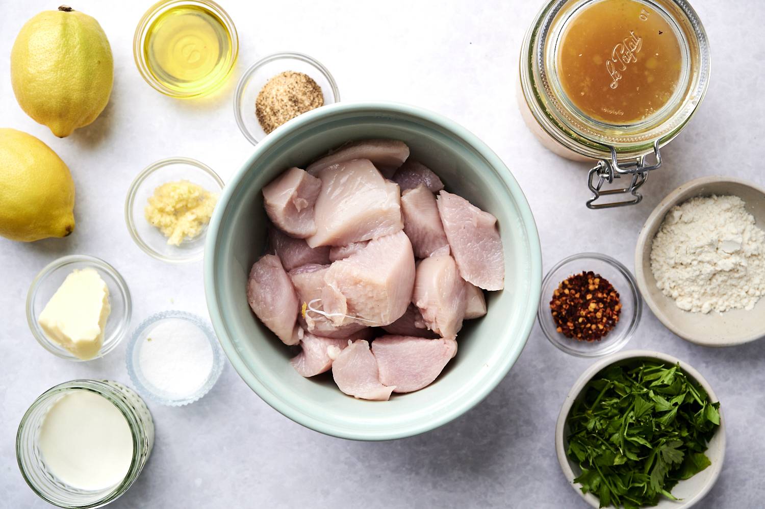 Bowl of raw chicken chunks surrounded by ingredients: lemons, garlic, oil, chicken broth, spices, butter, cream, flour, and fresh herbs on a white surface.