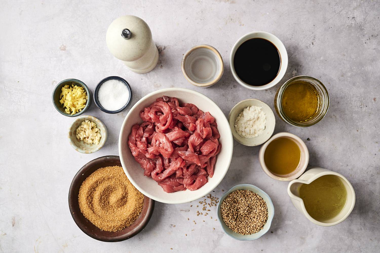 Raw beef strips in a bowl surrounded by ingredients like soy sauce, sesame seeds, garlic, and oil.