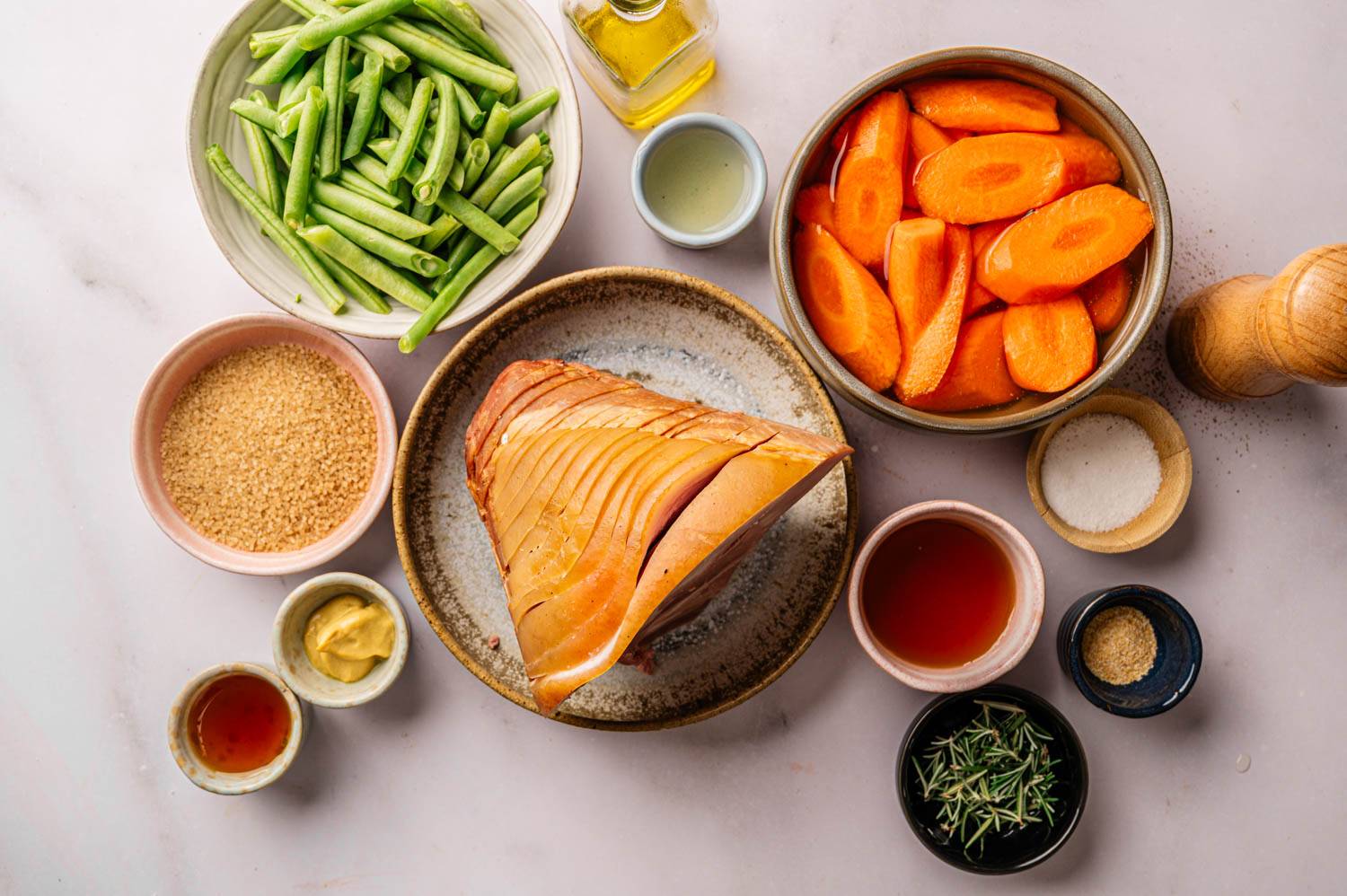 Ingredients for the roast are laid out on a marble surface: seasoned ham center, green beans, carrot slices, mustard, honey, salt, pepper, olive oil, brown sugar and rosemary.