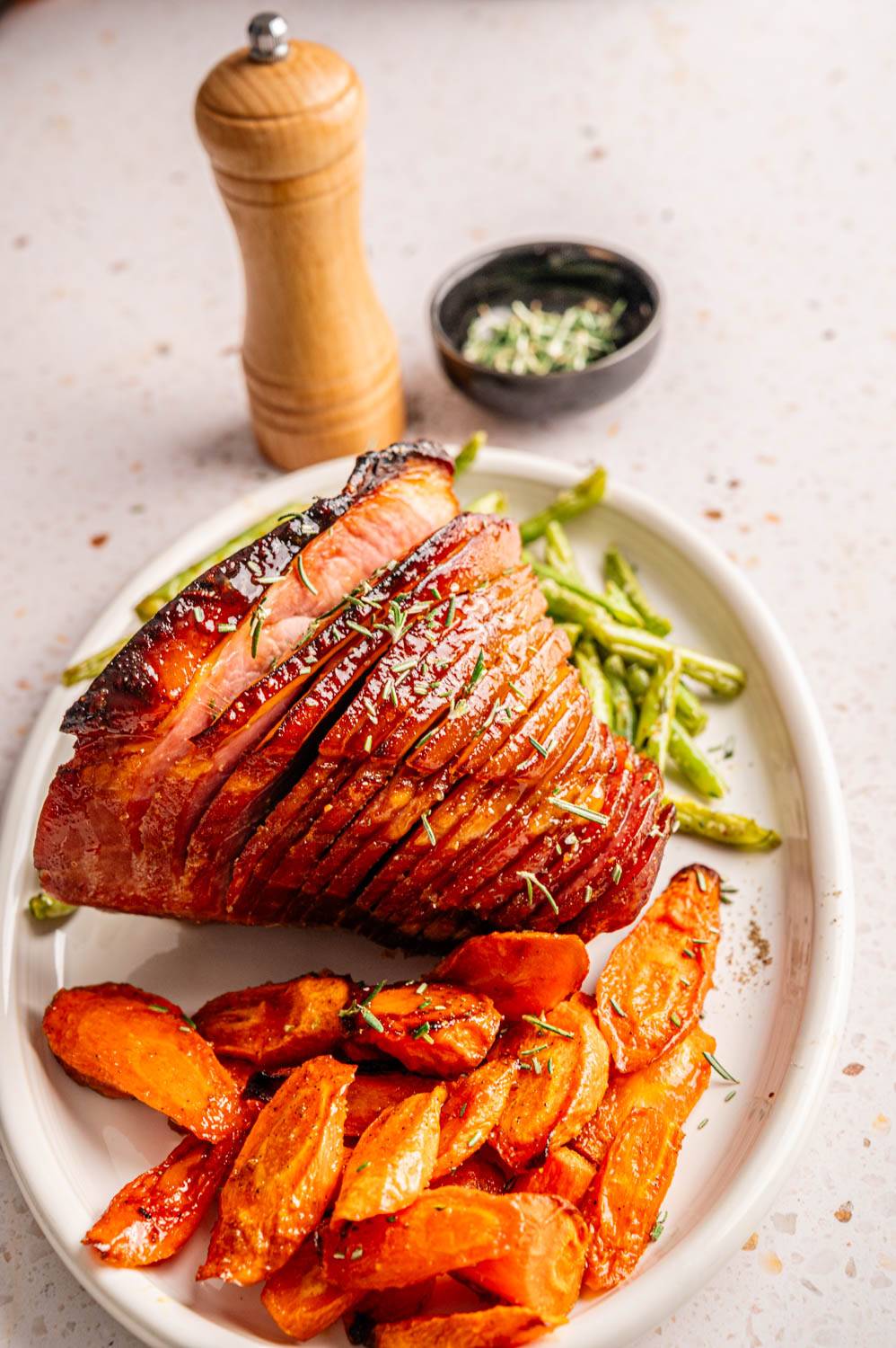 Sliced ham with rosemary on a white plate, served with roasted carrots and green beans. In the background are pepper grinders and herbs.