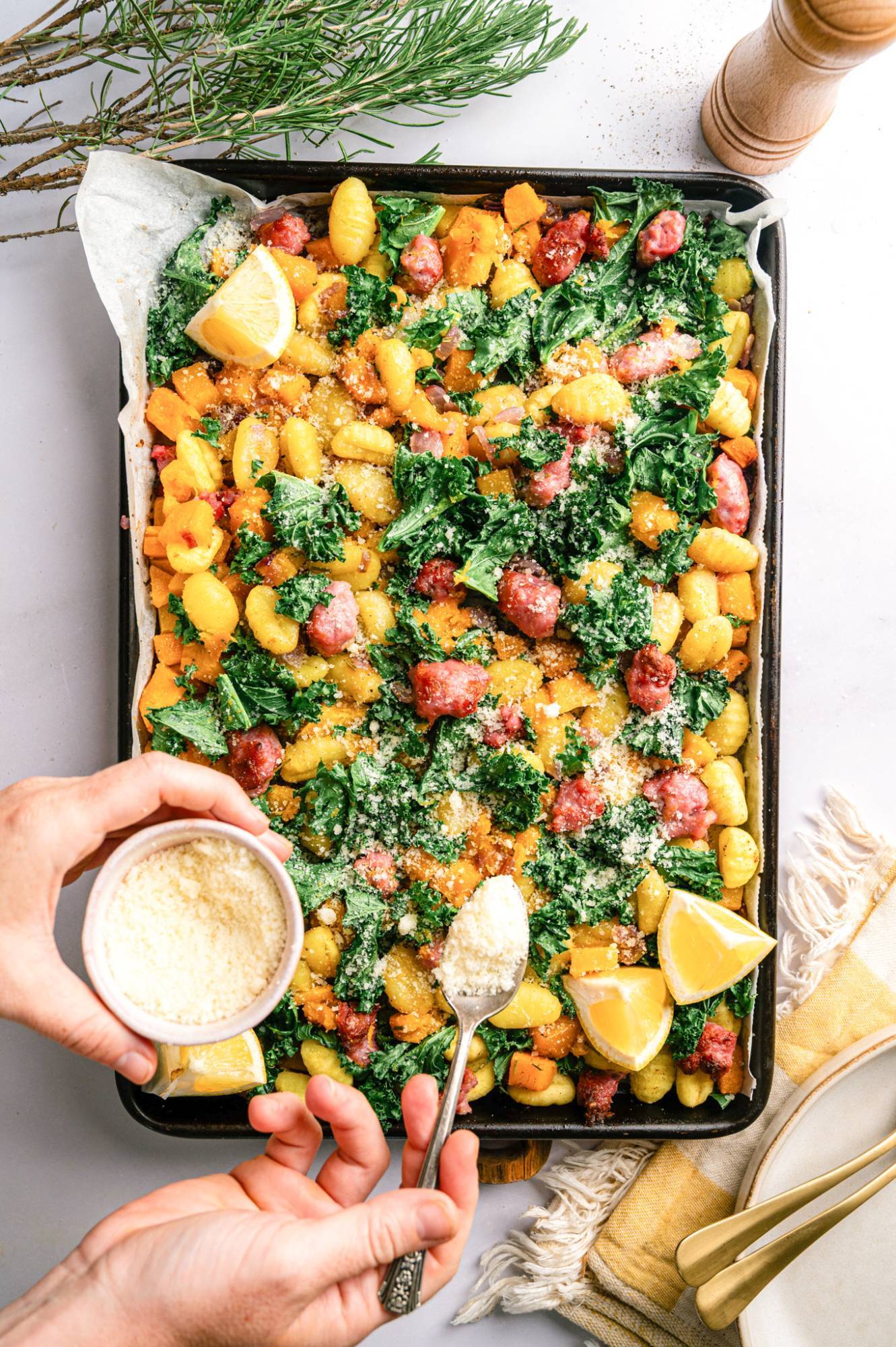 Parmesan cheese being sprinkled on a sheet pan with sausage, gnocchi, butternut squash, kale, and red onion.