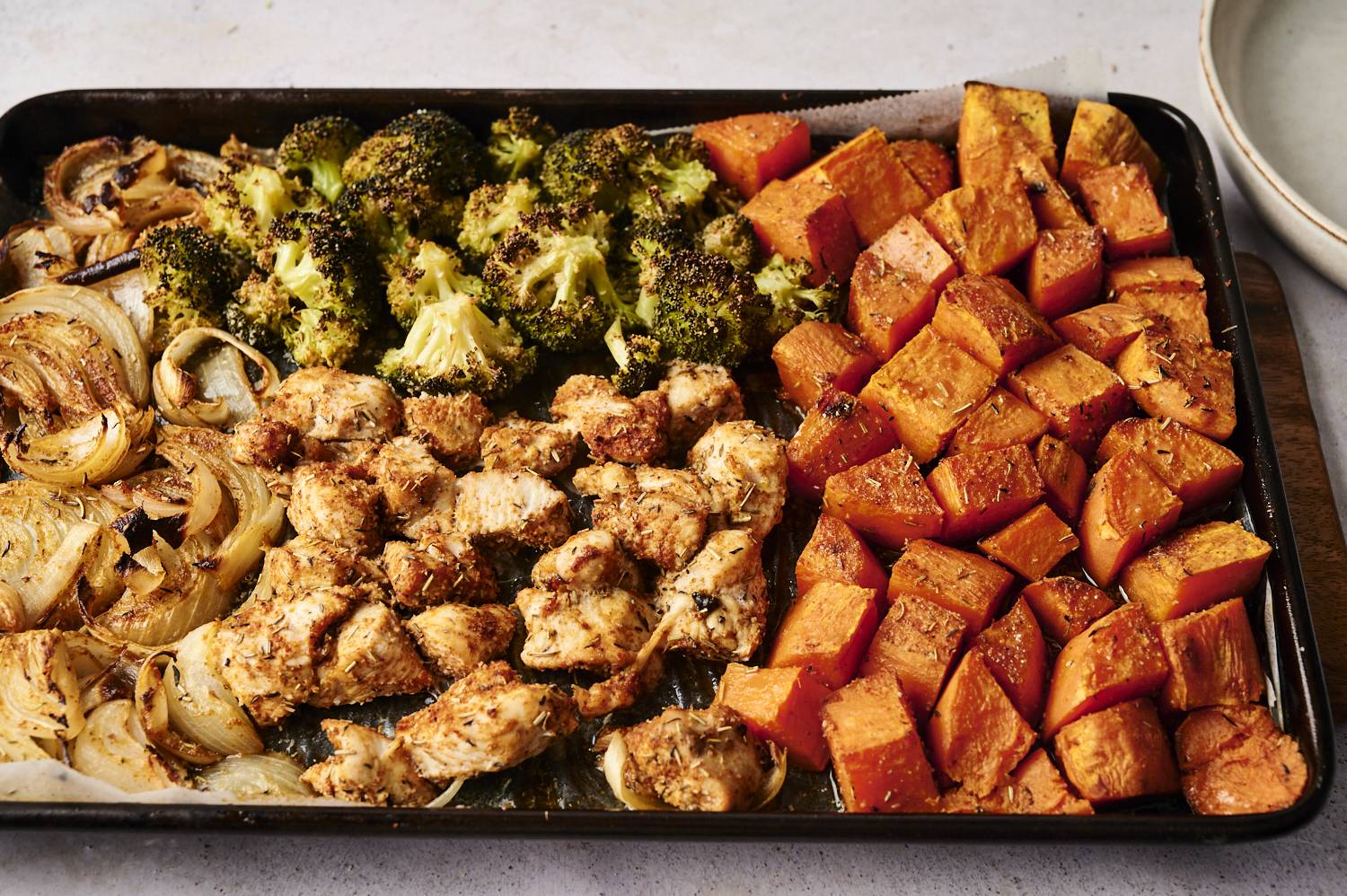 A sheet pan filled with roasted herb chicken, caramelized onions, roasted broccoli florets, and perfectly cooked sweet potato cubes. Each ingredient is arranged in separate sections on the pan, showcasing their golden, roasted textures.