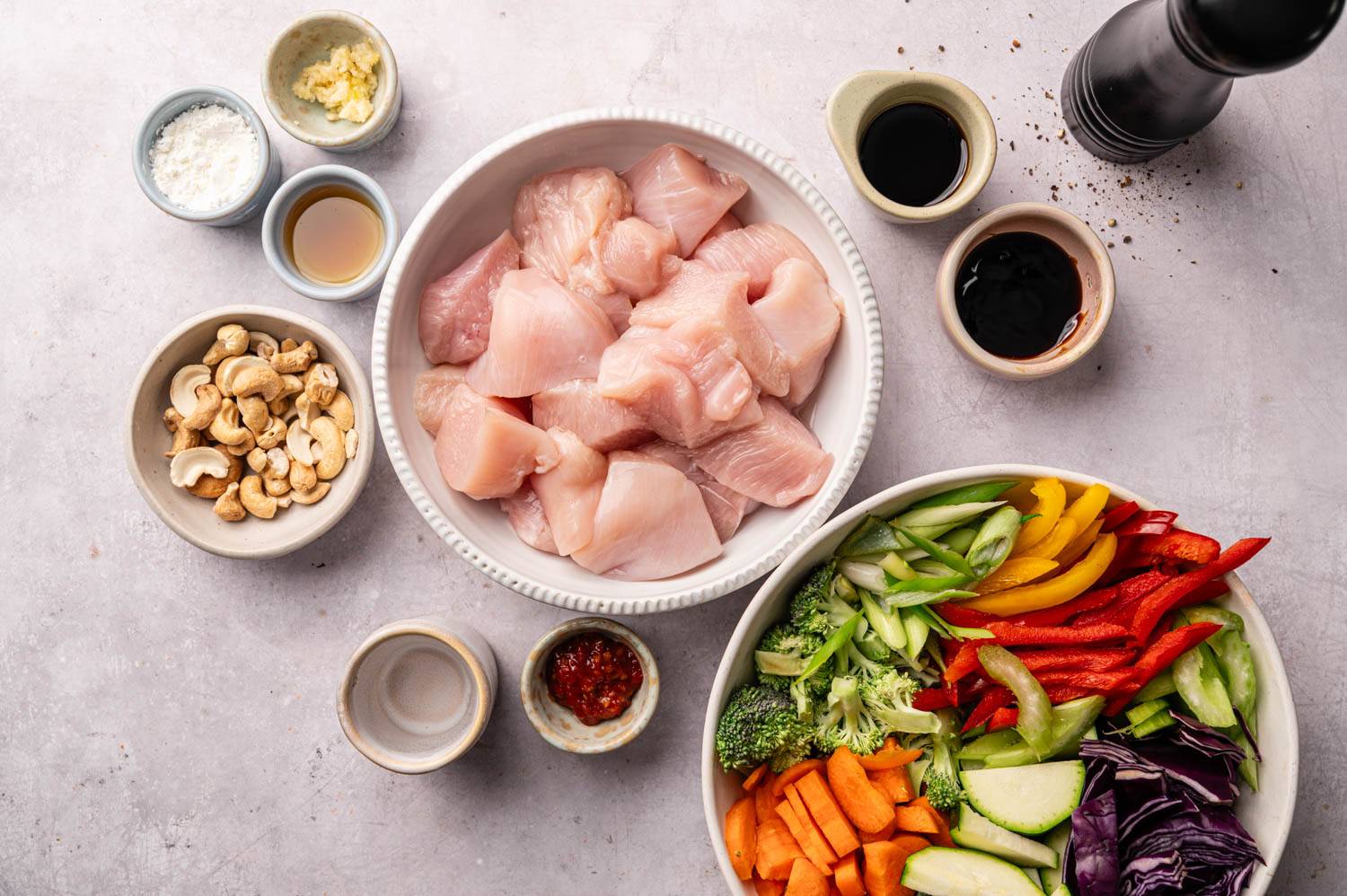 Raw chicken chunks in a bowl, surrounded by colorful sliced vegetables, sauces, cashews, and spices on a light countertop, ready for cooking.