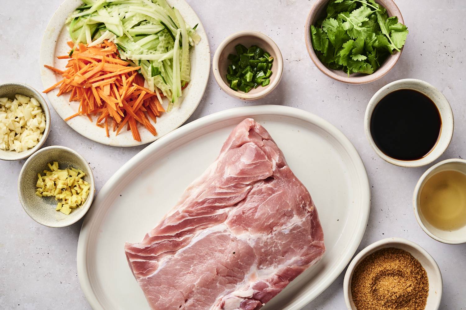A raw pork plate surrounded by fresh vegetables and seasonings including garlic, ginger, coriander and seasonings in small bowls.