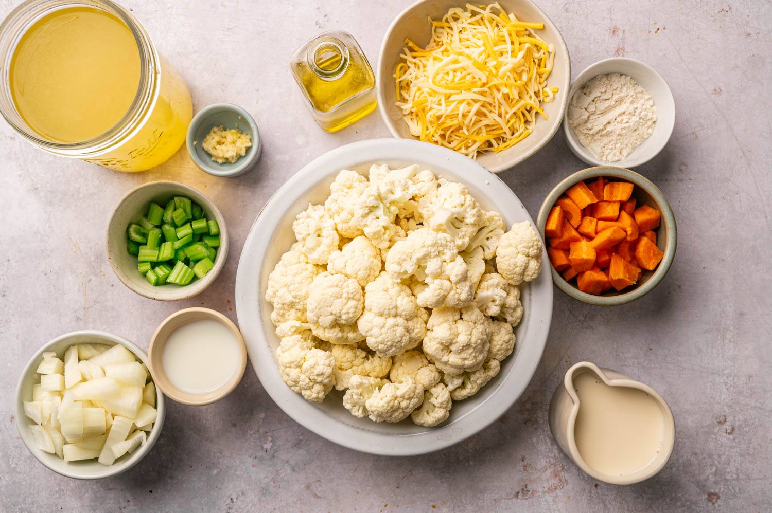 Top view of ingredients on countertop of cauliflower dish including cauliflower florets, grated cheese, chopped carrots, celery and onions, bowls of flour, broth, milk, garlic and olive oil.