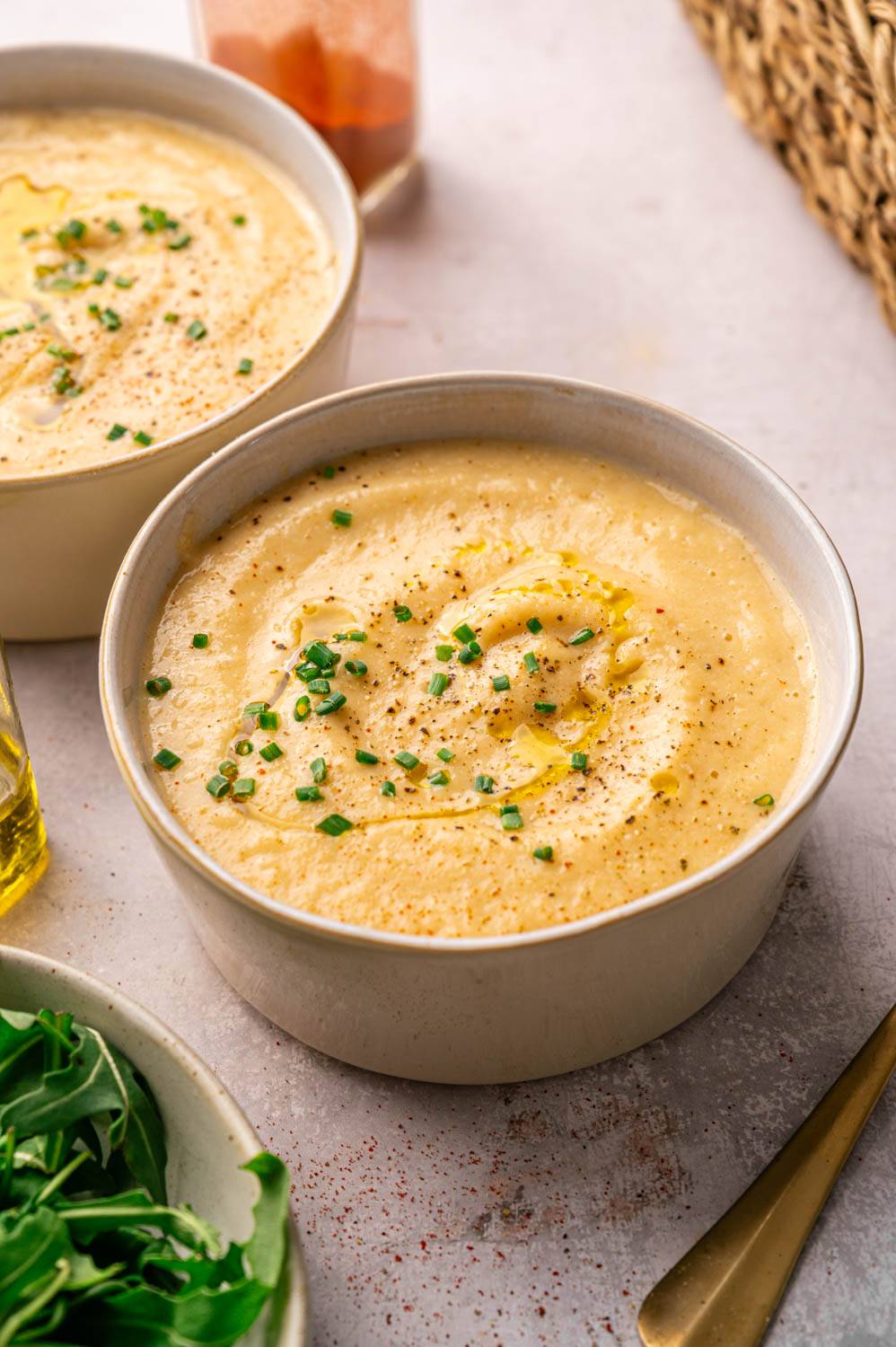 Cream soup in white bowl, garnished with chopped chives and black pepper. A hint of olive oil glistens on top. Fresh arugula and wicker baskets are nearby.