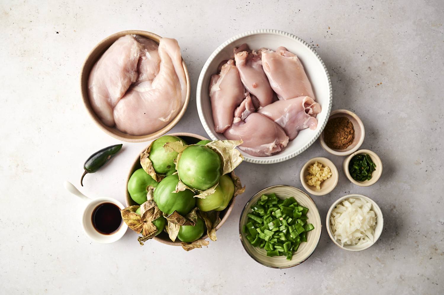Fresh ingredients for slow-cooker chicken in chile verde: raw chicken, tomatillos, green peppers, spices, and aromatics laid out on a light surface.