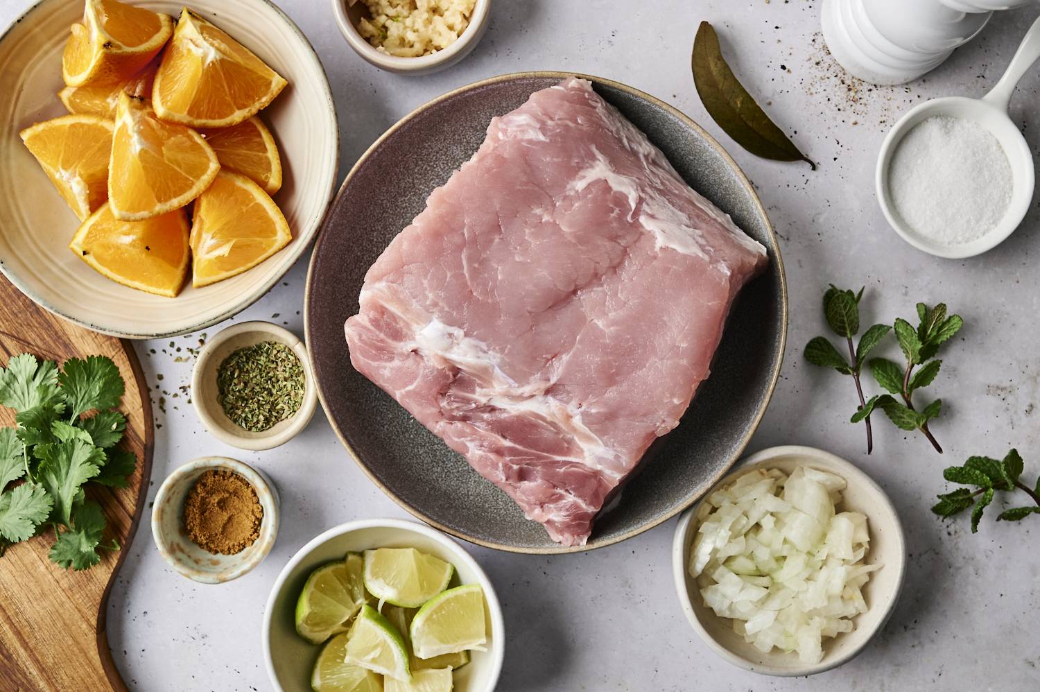 A collection of ingredients for slow-cooked garlic Cuban pork, including a pork roast, oranges, lime wedges, cilantro, onions, mint, and various spices like cumin, oregano, and bay leaves.