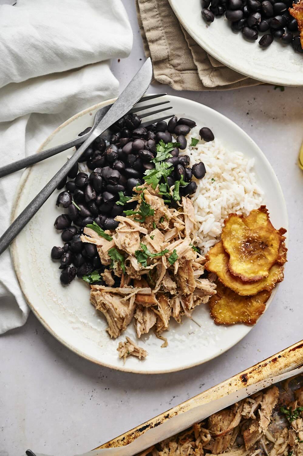 A plate with shredded garlic Cuban pork, black beans, cilantro, rice, and crispy plantains, with the pork laid over a bed of rice and garnished with fresh cilantro.