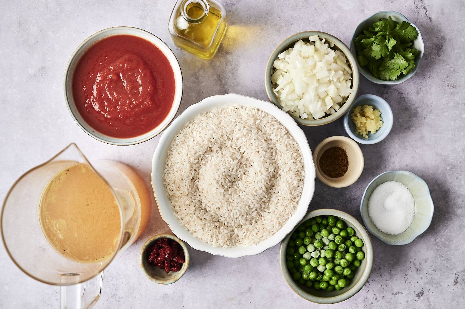 Overhead view of ingredients in slow cooker Mexican rice, including a large bowl of uncooked white rice surrounded by a bowl of tomato sauce, chopped onions, chopped garlic, frozen peas, tomato sauce, chicken soup, chicken stock, oil, chopped cilantro and spices, and spices on a light grey surface.